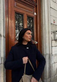 Kobieta w czarnym płaszczu trzyma beżową torebkę, stojąca obok ornamentowych drewnianych drzwi z żeliwnymi elementami, na tle kamiennej ściany.