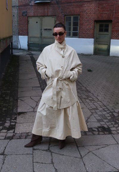 Mujer con abrigo trench beige extragrande y falda, con botas marrones hasta el tobillo, de pie en un patio adoquinado frente a edificios de ladrillo.