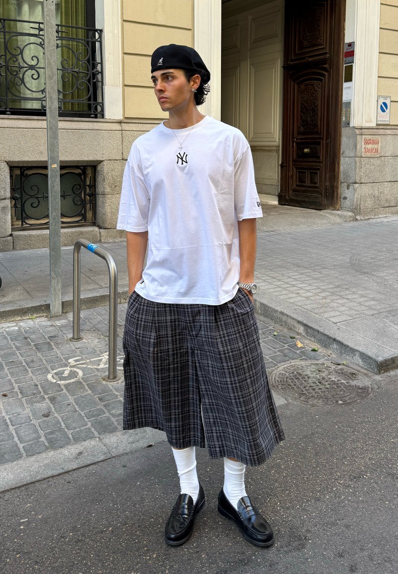 Jovem homem está de pé na calçada da cidade, usando um boné preto, uma camiseta branca oversized, calças largas xadrez, mocassins pretos e meias brancas.