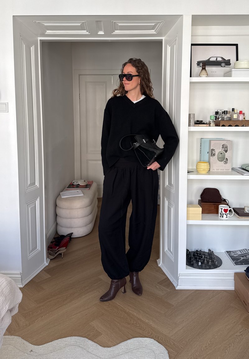 Femme portant un pull noir oversize, pantalon noir ample, bottines marron, lunettes de soleil foncées, tenant un sac à main noir dans l'embrasure d'une pièce moderne.