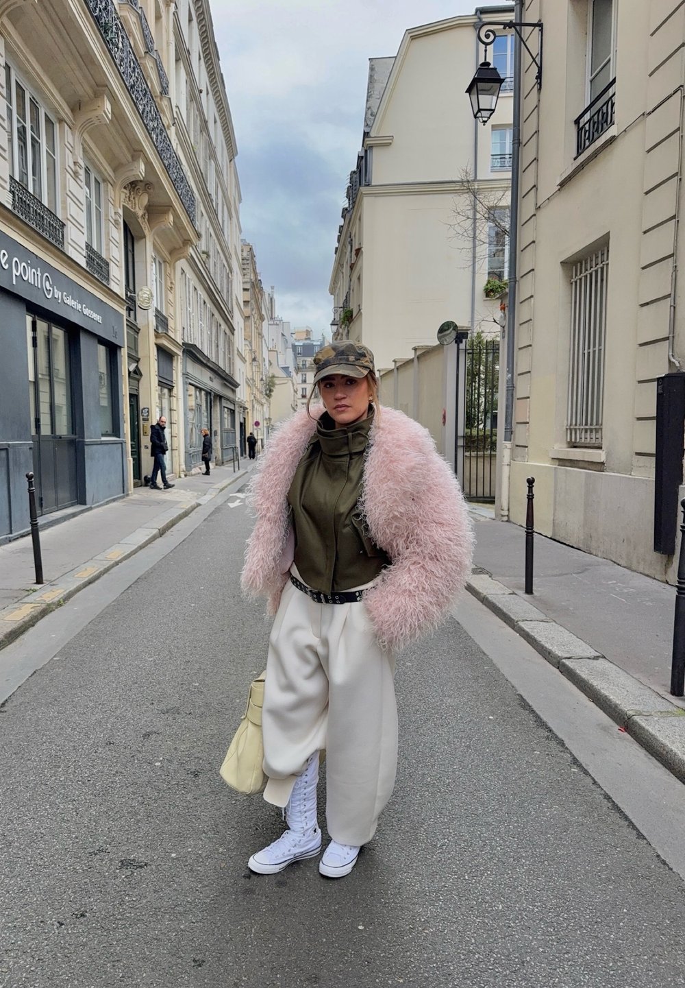 Femme portant une veste rose duveteuse, un haut vert olive, un pantalon large blanc, des baskets blanches, une casquette camouflage, debout dans une rue étroite de la ville avec des bâtiments de chaque côté.