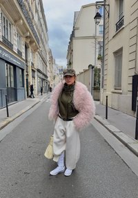 Femme portant une veste rose duveteuse, un haut vert olive, un pantalon large blanc, des baskets blanches, une casquette camouflage, debout dans une rue étroite de la ville avec des bâtiments de chaque côté.