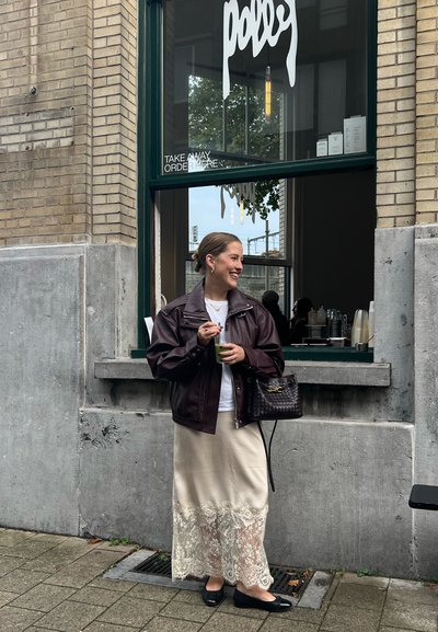 Chaqueta oversized de cuero marrón sobre una camisa blanca, combinada con una falda beige con acabado de encaje. Zapatos planos negros y un bolso tejido, sosteniendo una bebida.