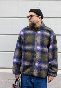 Fleecová bunda na zips v tlmenej zelenej a modrej farbe s károvaným vzorom s ružovými akcentmi. Výrazná textúra tkaniny a dizajn s vysokým golierom.