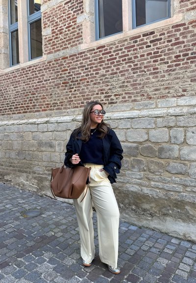 Bolso tote de cuero marrón, suéter azul marino, chaqueta negra, pantalones de pierna ancha color crema, gafas de sol con estampado, de pie contra una pared de piedra.