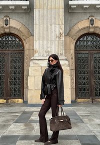 Czarna skórzana kurtka z wąskim pasem, dopasowana do ciemnobrunatnych spódnic w kształcie dzwonu. Trzyma ciemnobrunatną torbę z zamszu. Jednolity kolor, gładka tekstura.