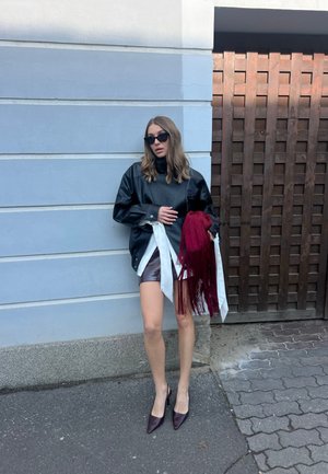 Mujer con chaqueta de cuero negra y gafas de sol, sosteniendo una bufanda roja con flecos, de pie junto a una pared azul pálido y una puerta de madera.