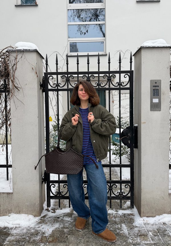 Ung kvinde i grøn jakke og blåstribet skjorte står ved en sort metalport i en snedækket bymiljø og holder en stor flettepose.