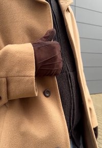 Παλτό από μείγμα καμήλας με καφέ σουέτ γάντι. Το παλτό έχει ορατό κουμπί και πέτο, σε συνδυασμό με μια υφασμάτινη μαύρη μπλούζα από κάτω.