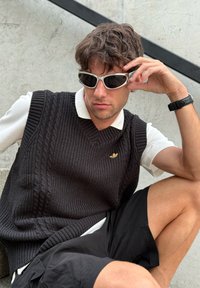 Colete de malha preto com design de ponto de cabos, camisa polo branca por baixo, calções pretos e óculos de sol brancos. Fundo suave e texturizado.