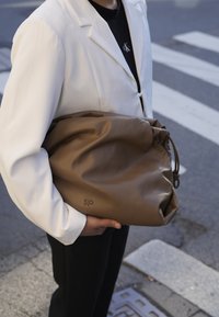 Καφέ δερμάτινη τσάντα με συγκεντρωμένο σχέδιο, ασφαλισμένη με κορδόνι. Διαθέτει διακριτό ανάγλυφο λογότυπο. Φοριέται κάτω από μία λευκή ζακέτα.