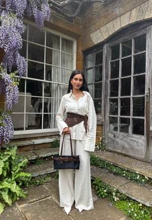 Mujer con conjunto blanco de rayas finas con cinturón marrón y bolso negro está junto a unas escaleras de piedra con flores moradas y puertas de vidrio detrás de ella.