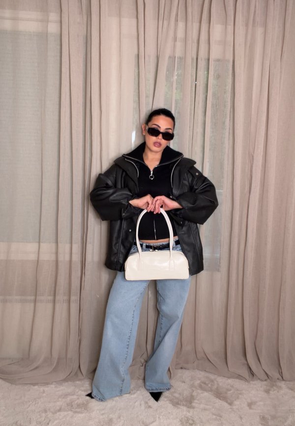 Femme portant une veste noire surdimensionnée, des lunettes de soleil et un jean bleu clair tient un sac à main blanc devant des rideaux beige sur un tapis clair.