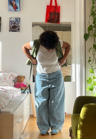 Jeans de corte holgado azul claro, camiseta blanca, chaleco verde y zapatos azules. Habitación luminosa con espejo, planta y oso de peluche sobre una cama.