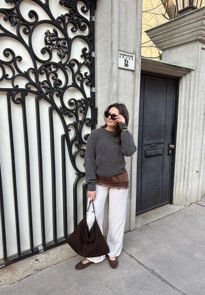 Mujer ajustándose las gafas de sol junto a una puerta de metal negro ornamentada, vistiendo un suéter gris, pantalones blancos, blusa interior marrón de encaje y sosteniendo un gran bolso marrón en la acera.