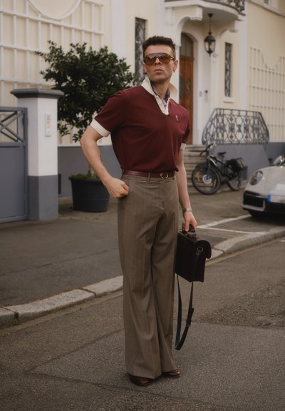 Polo burdeos con cuello blanco, pantalones beige a rayas, cinturón de cuero marrón y bolso; el modelo posa en la calle con gafas de sol.