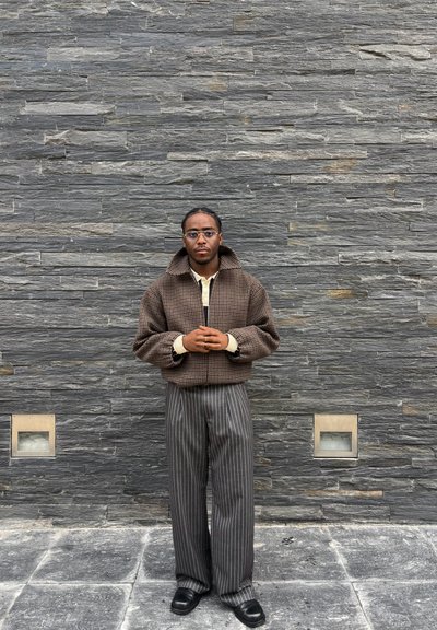 Joven con gafas que lleva una chaqueta marrón de cuadros y pantalones grises con rayas de espiguilla, de pie contra una pared de piedra gris, con las manos entrelazadas.