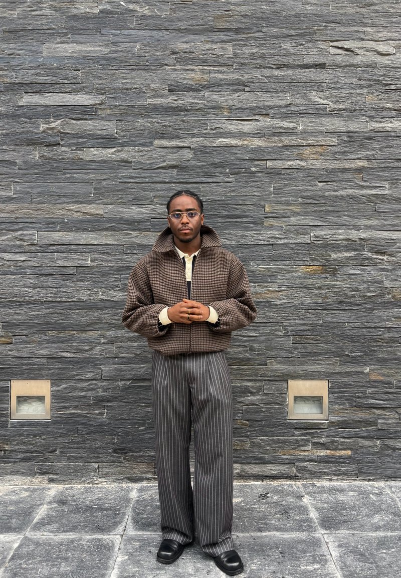 Noor mees prillidega, kes kannab pruunikat ruudulist jopet ja hallikat triibulist pükst, seisab halli kivimüüri taustal, käed kokkupandud.