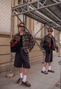 Model îmbrăcat cu o cămașă caramel cu carouri, un tricou negru, șorturi gri și pantofi cu șireturi maronii, purtând șosete albe. Ține o geantă neagră cu o eșarfă roșie.