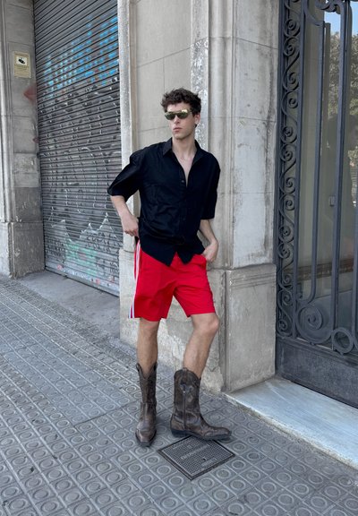 Camisa negra de manga corta, pantalones cortos rojos con rayas laterales y botas de vaquero de cuero marrón. De pie contra una pared de concreto texturizada.