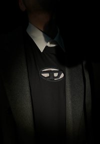 Svart genser med en svart brodert oval logo, lagd under en hvit skjorte med krage, delvis skjult av en mørk jakke.