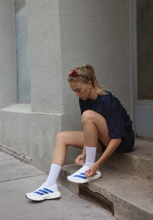 Scarpe da ginnastica bianche con strisce blu, indossate con calzini bianchi. Una persona sistema i lacci mentre è seduta su un gradino di cemento.