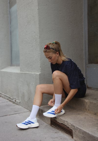 Zapatos deportivos blancos con rayas azules, combinados con calcetines blancos. Una persona ajusta los cordones mientras está sentada en un escalón de concreto.