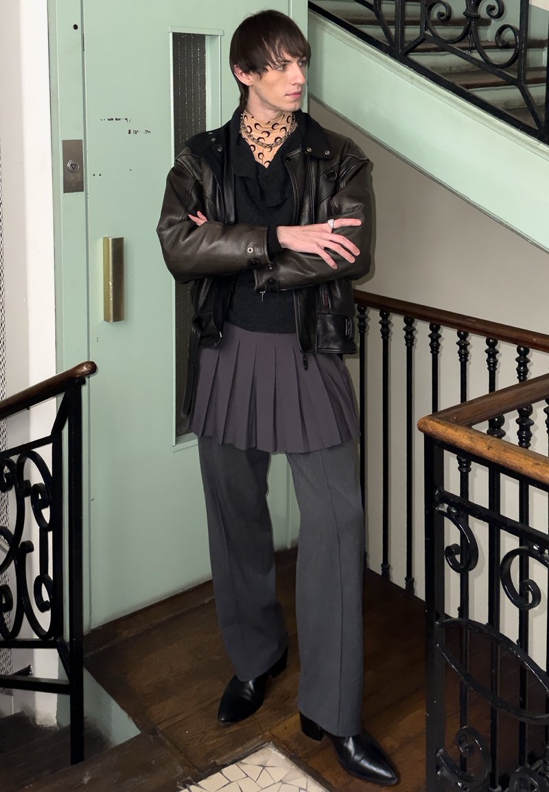 Personne debout dans un escalier, les bras croisés, portant une veste en cuir noire, un foulard à motifs autour du cou, une jupe plissée par-dessus un pantalon gris et des bottes noires.