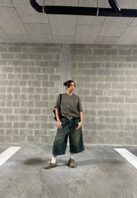 Camiseta recortada de color verde oliva, culottes de denim oversized con efecto desteñido, zapatos tipo slip-on beiges y un bolso negro. Fondo de concreto gris.