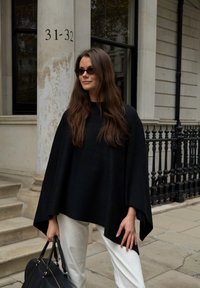 Fekete, laza szabású poncsó texturált anyagból, fehér nadrággal párosítva. Kiegészítve fekete kézitáskával és napszemüveggel.