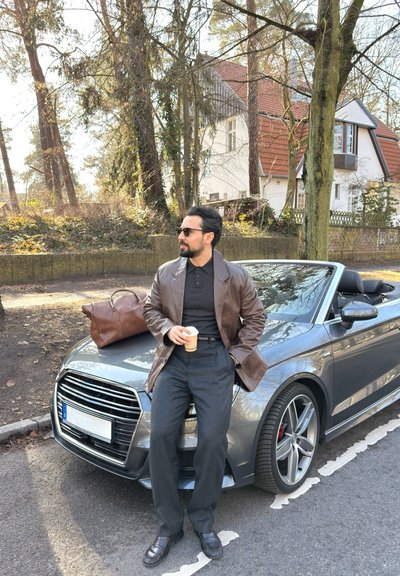 Hombre con gafas de sol y chaqueta de cuero marrón sosteniendo un café, apoyado en un coche descapotable plateado con una bolsa marrón sobre el capó, en una calle suburbana con árboles.