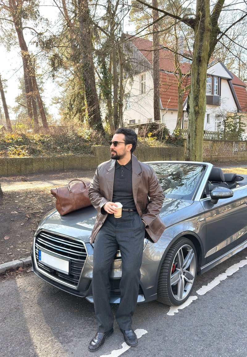 Άνδρας με γυαλιά ηλίου και καφέ δερμάτινο μπουφάν κρατώντας καφέ, στηριγμένος σε ασημί κάμπριο αυτοκίνητο με καφέ τσάντα στο καπό, προαστιακός δρόμος με δέντρα.