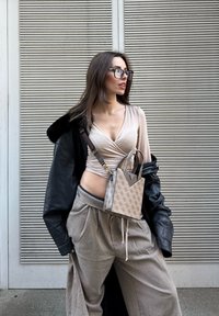 Jeune femme portant des lunettes, un haut beige enveloppant, un pantalon en laine oversized, et une veste en cuir noire posée sur les épaules, debout contre un mur rayé.