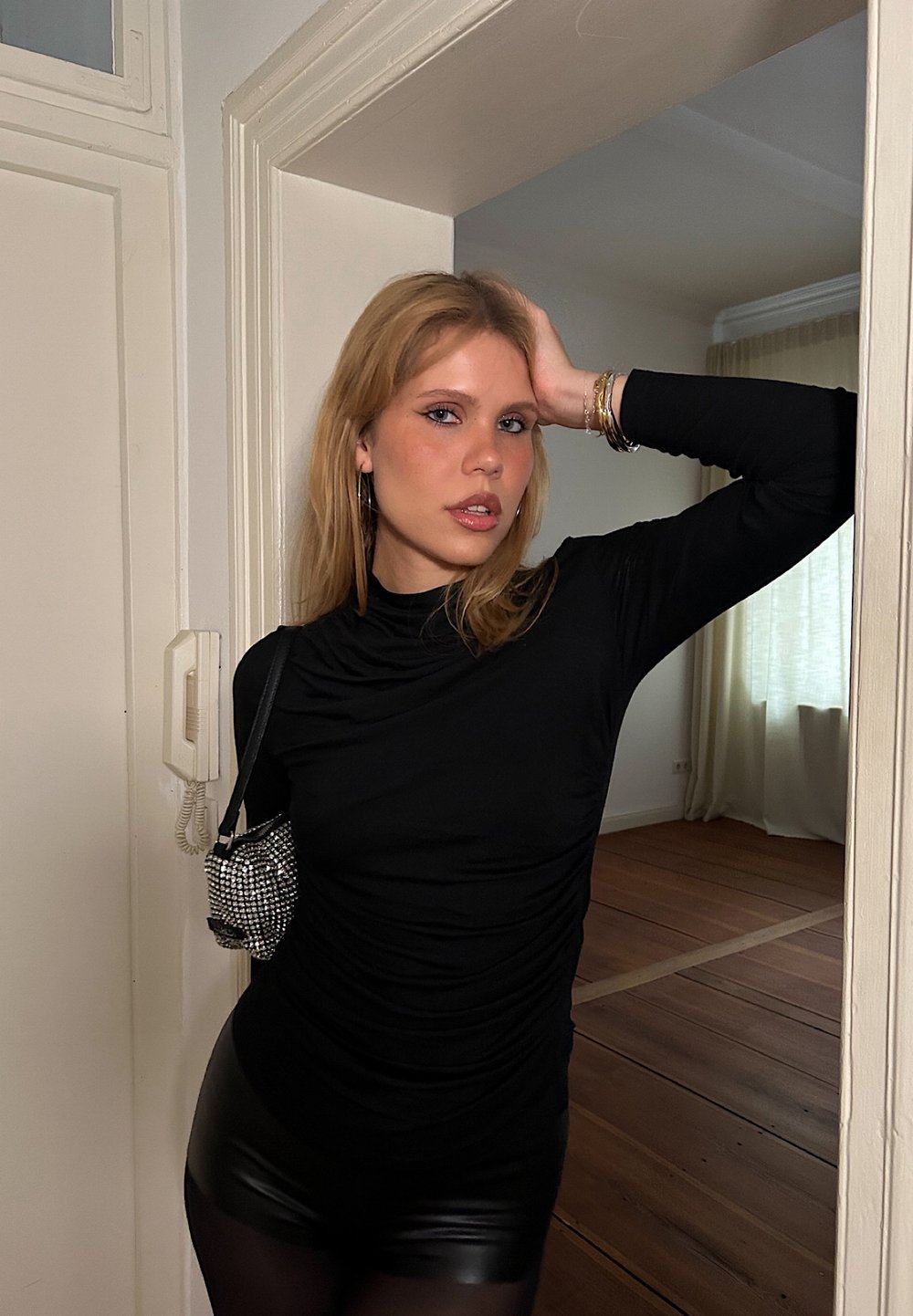 Μαύρο μακρυμάνικο τοπ με ρυτινωτή σχέδιο, συνδυασμένο με γυαλιστερές μαύρες σορτς. Μια μικρή, λαμπερή τσάντα κρέμεται από τον ώμο. Ο ξύλινος δάπεδο είναι ορατός.