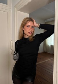 Μαύρο μακρυμάνικο τοπ με ρυτινωτή σχέδιο, συνδυασμένο με γυαλιστερές μαύρες σορτς. Μια μικρή, λαμπερή τσάντα κρέμεται από τον ώμο. Ο ξύλινος δάπεδο είναι ορατός.