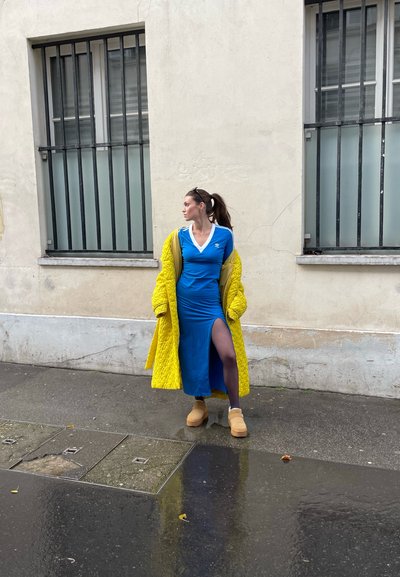 Vestido azul con una abertura lateral hasta el muslo, combinado con un abrigo largo acolchado amarillo y botas de plataforma beige. El escenario es una calle urbana con una ventana.