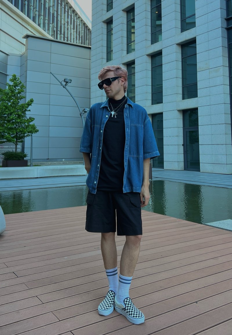 Jeune homme en lunettes de soleil portant une chemise bleue à manches courtes, un short noir et des chaussures à enfiler à carreaux, debout sur une terrasse en bois près de la piscine d'un bâtiment moderne.
