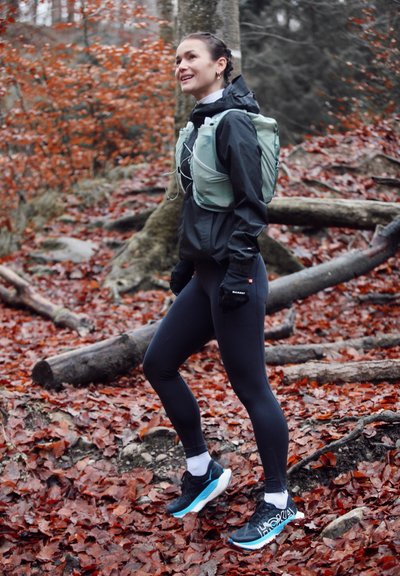 Corredor con chaqueta negra y mallas, calzando zapatillas de correr negras y azules. Mochila de hidratación verde claro en la espalda, con guantes visibles. Fondo de bosque.