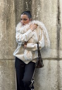 Femme appuyée contre un mur texturé portant une écharpe et un pull beiges en tissu duveteux, un pantalon noir avec des rayures blanches, tenant un sac à main marron foncé.