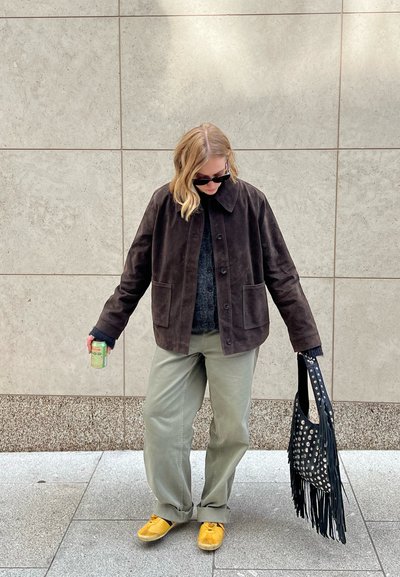Chaqueta de ante marrón con cuello, suéter gris, pantalones verde claro, zapatos amarillos y un bolso negro con flecos y tachuelas plateadas. Sosteniendo una lata.