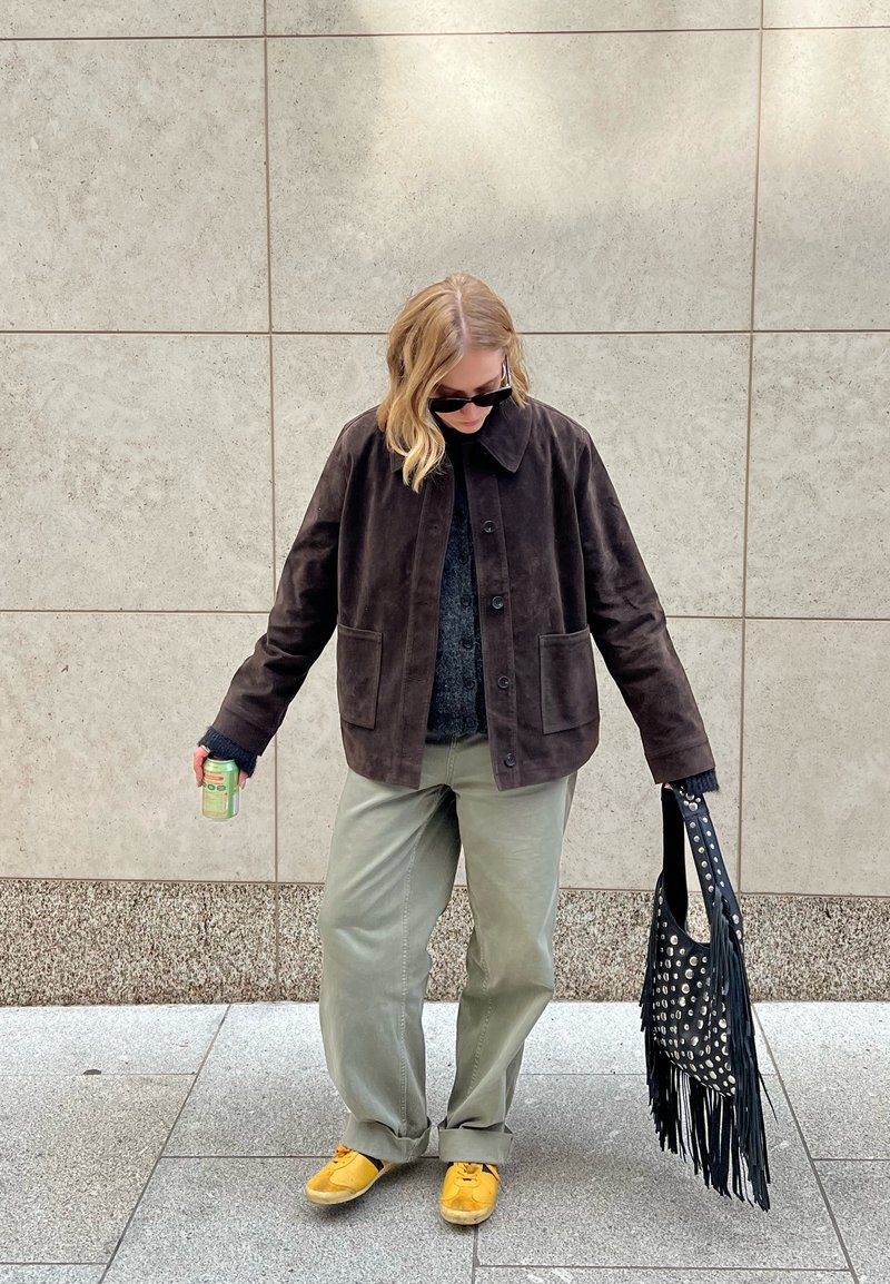 Giacca in suede marrone con colletto, maglione grigio, pantaloni verde chiaro, scarpe gialle e una borsa nera con frange e borchie argentate. Tiene una lattina.