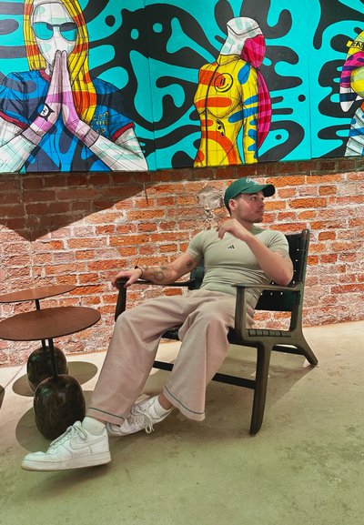 Silla de plástico negra con un diseño de listones; persona con una camiseta ajustada gris y pantalones de pierna ancha beige claro, zapatillas blancas y gorra verde.