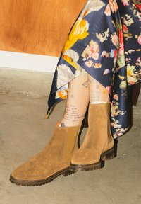 Personne portant des bottines en daim marron et une jupe à motif floral, assise sur une chaise sur un sol en béton.