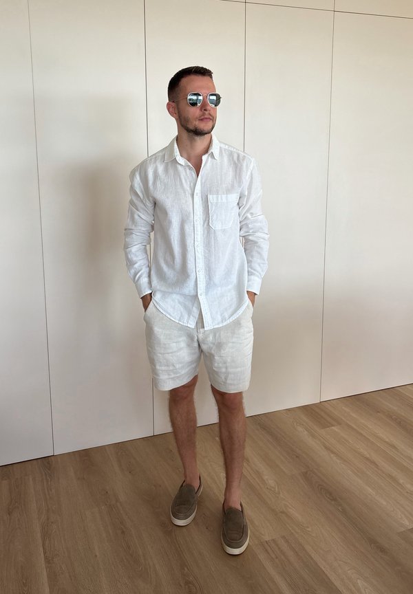 Homme portant une chemise blanche à manches longues, un short blanc, des chaussures marron sans lacets et des lunettes de soleil, debout à l'intérieur sur un sol en bois contre un mur beige.