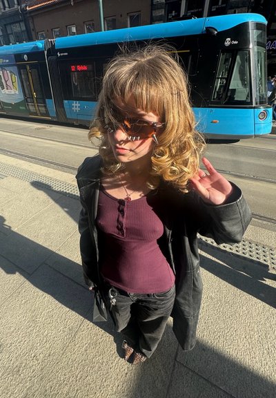 Una persona que lleva una camiseta de canalé morada y pantalones negros, una chaqueta de cuero negra de gran tamaño y gafas de sol con pinchos, está de pie al aire libre con un tranvía detrás.