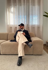 Μπεζ καναπές από ύφασμα με λεία υφή. Άτομο καθισμένο με τα πόδια σταυρωμένα, φορώντας σκούρο σακάκι, ανοιχτόχρωμο παντελόνι και μαύρα παπούτσια, κρατώντας μια κούπα.