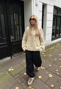 Jersey beige de punto grueso, pantalones anchos negros y zapatos marrones sin cordones. De fondo se ve un edificio con puertas negras y hojas caídas.