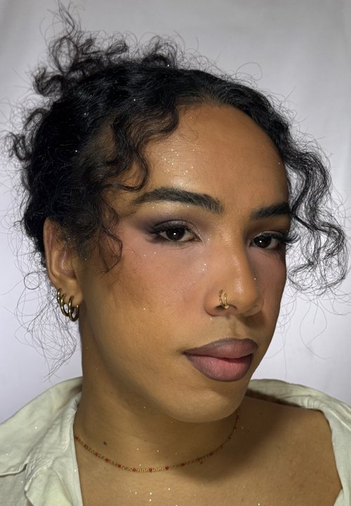 Personne avec un chignon aux cheveux bouclés, portant trois boucles d’oreilles anneaux en or à l'oreille gauche, un anneau de nez en or, un maquillage subtil et un délicat collier rouge perlé.