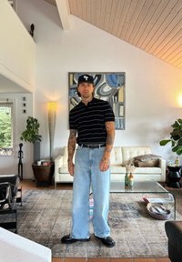 Hombre con tatuajes que lleva una camisa negra a rayas, pantalones anchos de color azul claro y zapatos negros, de pie en una habitación moderna con plantas y obras de arte.