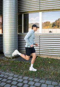 Vīrietis sporta tērpā skrien blakus rūpnieciska stila metāla ēkai ar lielu logu, kurā atspoguļojas zaļā ainava.